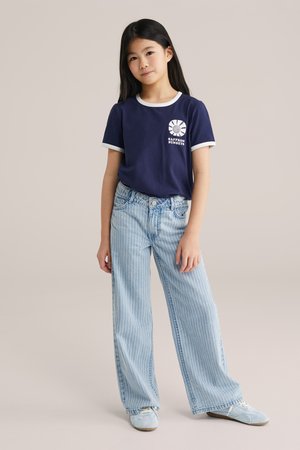 Joven niña de pie con camiseta azul marino "Saffron Sunsets", vaqueros de pernera ancha azul claro con rayas finas y zapatillas deportivas azul claro sobre fondo liso.