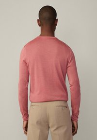 Rosa Pullover mit langen Ärmeln, gerippter Textur und Ellbogenflicken; kombiniert mit hellbeigen Hosen. Der Pullover hat einen runden Ausschnitt.