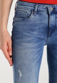 Jeans de mezclilla azul desgastados con un diseño ajustado, que presentan una ligera área deshilachada en el muslo y un estilo clásico de cinco bolsillos.