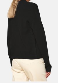 Mustat neulotut villapaidat, joissa on pyöreä kaula-aukko ja pitkät hihat, rento istuvuus ja sivuhalkiot. Pääkallio vaaleiden housujen päällä.