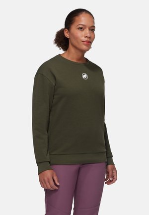 Kvinde iført en olivengrøn sweatshirt med et lille hvidt logo på brystet og lilla bukser, stående mod en neutral baggrund.