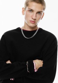 Maglione nero a maglia con scollatura rotonda, indossato sopra un top bordeaux. Collana in argento e bracciale con dettaglio a chiusura sul maniche.
