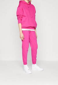 Felpa rosa brillante con tasca a marsupio, abbinata a pantaloni jogger coordinati. Presenta polsini a coste e una maglietta bianca sotto. Indossata con scarpe da ginnastica bianche.