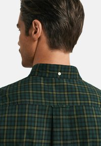 Camicia a quadretti verde con linee gialle e nere, un bottone al colletto e un dettaglio a plissettatura sul retro. Tessuto strutturato, design casual.