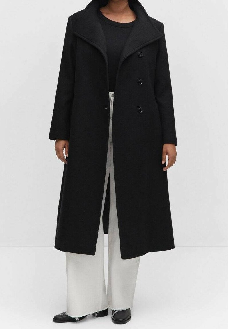 Cappotto lungo nero con colletto alto, dotato di tre bottoni frontali, realizzato in un tessuto simile alla lana. Indossato sopra una maglietta nera e pantaloni grigio chiaro.