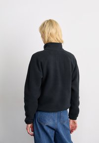 Persona con capelli biondi corti che indossa una giacca in pile nera e jeans blu, di fronte a uno sfondo bianco semplice.