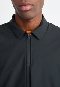 Sort hættetrøje med lynlås og krave samt en orange inderside. Glat overflade og tætsiddende design, der har en central lynlås og ingen synlige mønstre.