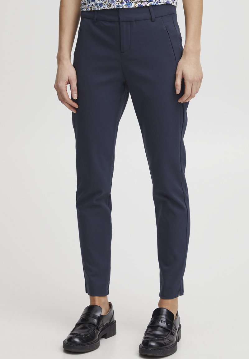 Marineblauwe slim-fit broek van gladde stof. Heeft een vlakke voorkant, riemlussen en zijzakken. Gecombineerd met zwarte instapschoenen.