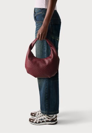 Burgundy vævet lædertaske med et rundet design og kort håndtag, parret med højtaljede blå jeans og brune sneakers.