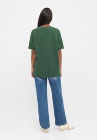 Groene katoenen t-shirt met korte mouwen, relaxte pasvorm en ronde hals. Draagt men met blauwe wijde spijkerbroeken en lichtgekleurde schoenen.