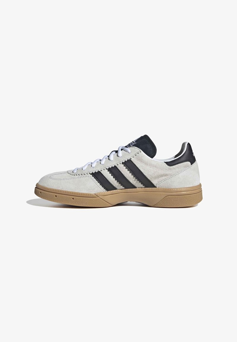 Chaussure en daim blanc avec des accents noirs, trois bandes noires sur le côté, bout rond, lacets blancs et semelle en caoutchouc gomme.