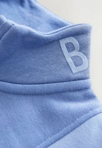 Prim-plan al unui guler înalt din material albastru deschis, cu litera mare „B” cusută pe lateral.