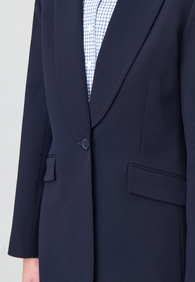 Blazer en tissu marine avec une texture lisse, revers pointus, fermeture à un bouton et deux poches avant. Porté par-dessus une chemise à carreaux bleue.