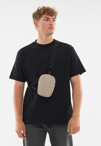 Borsa a tracolla in beige chiaro, forma rettangolare con texture liscia. Dotata di tracolla nera regolabile e chiusura con zip per un accesso sicuro.