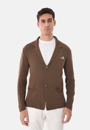 Uomo che indossa un blazer marrone con trama, con due bottoni, una camicia bianca sotto, pantaloni bianchi e un fazzoletto da taschino con motivo.