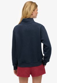 Sudadera de color azul marino con cuello alto, puños y cintura acanalados. Llevada con pantalones cortos rojos, presenta un parche cuadrado de tela en la parte posterior.