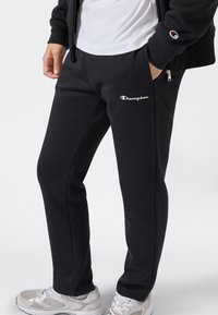 Uomo che indossa pantaloni tuta neri Champion con logo bianco sulla coscia, scarpe da ginnastica bianche, t-shirt bianca e felpa nera con zip e logo sulla manica.