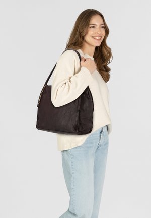 Borsa a spalla in pelle marrone con una texture liscia, forma strutturata e borchie decorative lungo la tracolla. Indossata da una persona.