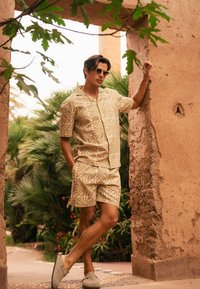 Korte mouwen shirt en shorts set in lichtbeige met een witte bladdessin. Draagt grijze instap schoenen en een zonnebril. Groene bladeren op de achtergrond.