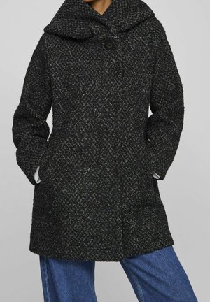 Kvinde iført en mørk tekstureret uldfrakke med stor krave og knap, hænderne i lommerne, kombineret med blå jeans mod ensfarvet baggrund.