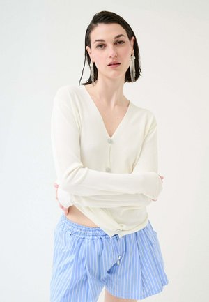 Giovane donna con capelli corti e scuri, indossa un cardigan bianco con bottoni e pantaloncini a righe blu, posa con le braccia incrociate su uno sfondo neutro.