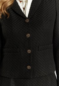 Blazer nero con texture, caratterizzato da un colletto a punte, cinque bottoni decorativi con design floreali e due tasche frontali.