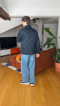 Νέος άνδρας με μαύρο καπιτονέ μπουφάν και φαρδιά τζιν στέκεται σε ένα μοντέρνο καθιστικό με καφέ καναπέ και μεγάλο γλαστρικό φυτό.