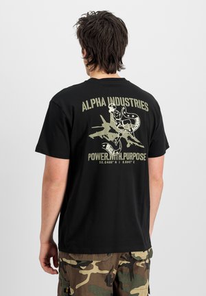Persoon die van achteren te zien is, draagt een zwart Alpha Industries T-shirt met een afbeelding van een gevechtsvliegtuig en pin-up meisje, gecombineerd met camouflage cargobroeken.