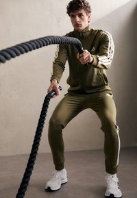 Olíva zöld sportos tréningruha fehér három csíkkal, fekete harci kötél és fehér sportcipő, semleges háttér előtt.