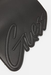 Černý kožený doplněk s vystouplým logem „Guess“, hladkou texturou a kontrastním prošitím podél okrajů. Celkový tvar je obdélníkový.