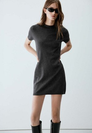 Femme portant une robe courte cintrée gris charbon, des bottes noires hautes jusqu'aux genoux et des lunettes de soleil noires, les mains derrière le dos.