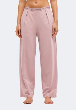 Femme portant un pantalon de détente large rose clair avec une taille élastique estampillée Lacoste, debout pieds nus devant un fond uni.