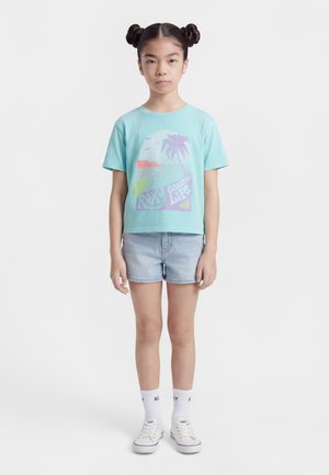 Junges Mädchen mit Doppelknoten-Frisur, das ein türkisfarbenes T-Shirt mit Grafik, helle Jeansshorts, weiße Socken und weiße Turnschuhe trägt und stillsteht.