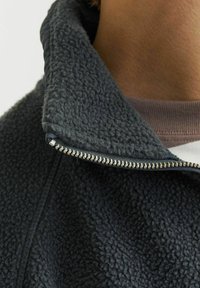 Fleece jakke med høj krave, mørkegrå farve, tekstureret stof og en sølvmetal lynlås foran.
