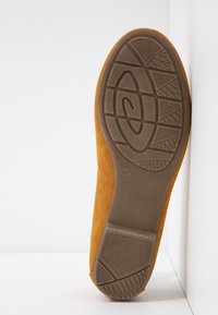 Chaussure en daim couleur fauve avec une semelle en caoutchouc texturée présentant un motif ondulé et des lignes d'adhérence. La chaussure a un bout arrondi et une finition lisse.