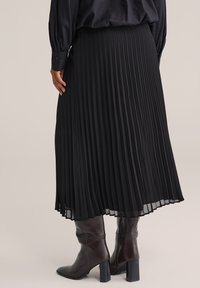 Gonna midi plissettata nera indossata con stivali in pelle scuri con tacco e una blusa a maniche lunghe, vista da dietro.