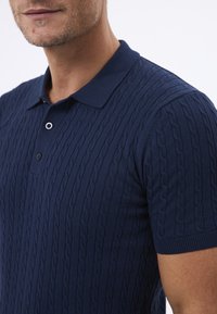 Mørkeblå polo-shirt med et struktureret kabelstrik mønster, korte ærmer og en klassisk krave. Enkel knap ved halsudskæringen.