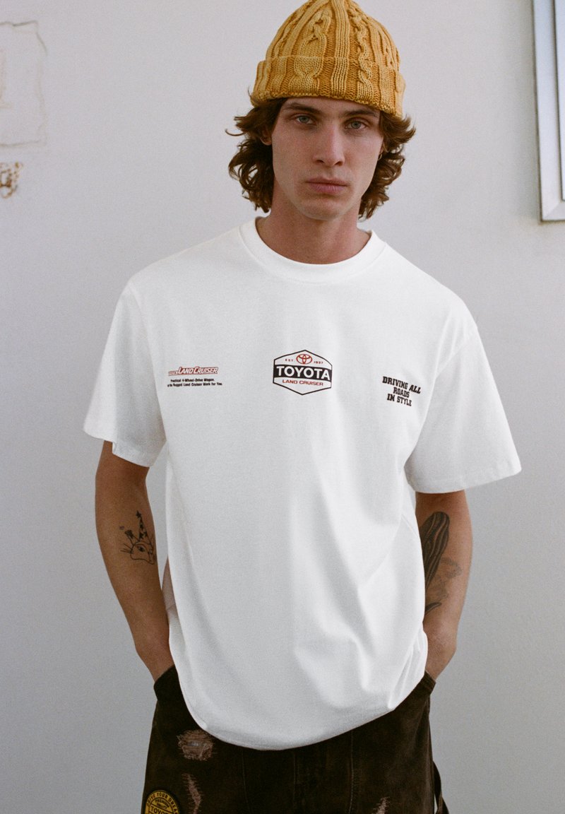 Biała bawełniana koszulka z logo i napisem "TOYOTA" na przodzie. Okrągły dekolt, luźny krój. Noszona z ciemnymi wzorzystymi spodniami i żółtą dzianą czapką.