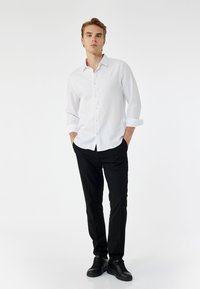 Camisa blanca de botones con mangas remangadas, combinada con pantalones negros ajustados y zapatillas negras. Tejido suave y diseño limpio.