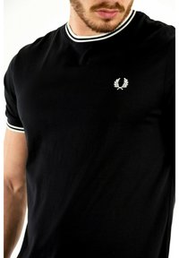 Sort kortærmet t-shirt lavet af bomuld, med en ribbet krave med hvide striber og et hvidt broderet logo på brystet.
