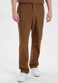 Brune cargo bukser med elastisk talje, to store frontlommer og en afslappet pasform, parret med hvide sneakers på en neutral baggrund.