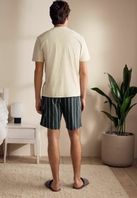 Helles beige T-Shirt mit kurzen Ärmeln, gestreifte dunkle Shorts und graue Hausschuhe. Der Hintergrund zeigt eine Topfpflanze und einen Nachttisch mit einer Lampe.