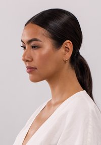 Goldene Creolen werden von einer Frau mit langen, dunklen, zurückgebundenen Haaren getragen. Sie hat einen natürlichen Make-up-Look und trägt ein weißes Oberteil mit V-Ausschnitt.