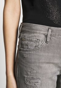 Jeans grises desgastados con dobladillos deshilachados y bolsillos. La tela es de mezclilla, presenta un cierre de botón y cremallera, con un ligero brillo.