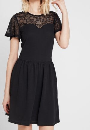 Woman wearing a black dress with a lace yoke and flutter sleeves, fitted bodice, and a gathered skirt, posing with one hand near her head.
