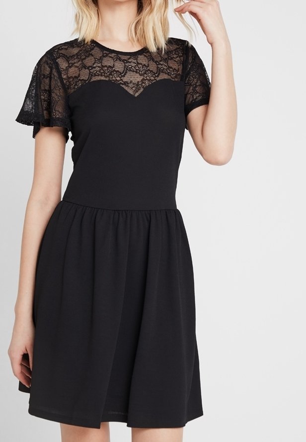 Femme portant une robe noire avec un empiècement en dentelle et des manches légères, un corsage ajusté et une jupe froncée, posant avec une main près de sa tête.