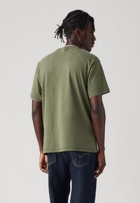 Tricou din bumbac de culoare verde măslin, cu mâneci scurte, croială lejeră, decolteu rotund și un tiv care ajunge până la șolduri, asortat cu blugi din denim închis.