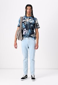 Kortermet blå blomstret skjorte, lyseblå jeans, sorte joggesko med hvite detaljer, bærer en grå crossover-veske.