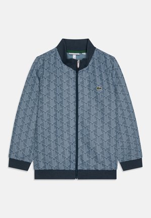 Veste zippée bleu et marine, avec un motif géométrique sur l'ensemble. Col et poignets côtelés, avec un petit accent logo vert.