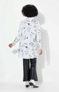 Chemise blanche avec des motifs graphiques noirs, manches longues et col. Associée à un pantalon évasé noir brillant et des bottes métalliques argentées.